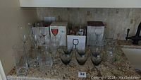 Assorted wine glasses, tumblers, champagne glasses, mugs on granite countertop, some in original boxes.