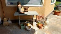 Overall view of stone table with wrought iron legs and assorted ceramic pots, metal bird bath, and decorative items surrounding it.