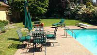 Full view of round glass-top table, four chairs, chaise lounge, side table, umbrella and stand by pool