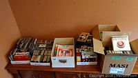 Multiple boxes containing DVDs, CDs, cassette tapes and VHS tapes arranged on a wooden surface