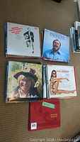 Top view of assorted vinyl records on carpet, including albums by Neil Diamond, Luciano Pavarotti, John Denver, and Barbra Streisand, with a red vinyl record sleeve visible