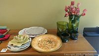 Full view of assorted vintage china plates, green glass vases, small dark green glass items, and decorative pieces on wooden table.