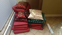 Stack of assorted patio cushions and decorative throw pillows, showing red striped cushions, green striped cushion, cream floral pillow, and dark red floral pillows.