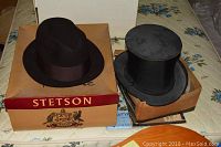 Photo showing black Stetson fedora hat with brown ribbon on original Stetson box next to a black top hat in worn box.