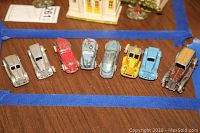 Photo showing eight vintage toy cars lined up on a wooden surface, displaying various paint colors and styles from silver to brown, including a yellow utility vehicle.