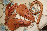 Photo showing the two brown leather toy holsters decorated with colorful studs, with one holster holding several red plastic toy cap guns, plus a braided leather whip coiled beside them on a floral fabric surface.