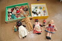 Top-down view of two boxes containing various traditional dolls in colorful outfits, plus five dolls laid out including a straw doll and fabric dolls with aprons.