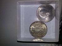 Two coins placed in a small white box showing the obverse of 1965 foreign coin and reverse of the Peace Dollar.