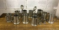 14 assorted pewter beer steins on wooden surface, some with hinged lids and clear bottoms.