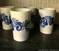 Four cream-colored pottery cups with blue swirl and floral design, approximately 4 inches tall, placed on black surface.