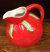 Front angle view of the red ceramic jug with green leaf accents, showing the rounded body, handle, and spout.