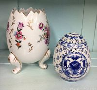 Pair of decorative egg-shaped china dishes shown side by side on a shelf, larger floral vintage egg with tripod legs on left, smaller blue and white Bombay egg dish on right.