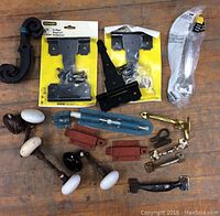 Photo shows assorted hardware on wood surface including new Stanley hinges, metal handles, brass latch, and porcelain door knobs.