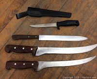 Four knives arranged on wood surface, showing blades and handles, including a small fixed blade knife with black handle and sheath, a long wooden handled straight blade, and two long wooden handled curved blades.