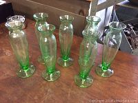 Photo showing seven green glass vases arranged on a table, each about 6 inches tall with flared tops and wide bases.