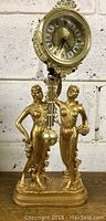 Front view of gold-tone resin clock with two standing female figures and pendulum under clock face
