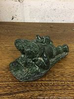 Green ram sculpture shown on wooden surface, angled view showing detailed curling horns and texture of fur.