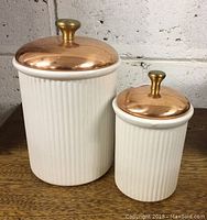 Two white ceramic canisters with vertical ribbing and copper lids with brass knobs on a wooden surface against a wall, larger canister approx. 8in tall