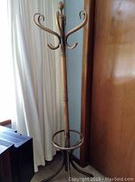 Full view of the bamboo coat tree rack showing the top hooks, upright post, circular stabilizing ring, and tripod base against a background of a window with curtains and wooden door.