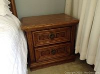 Front view of wooden night table positioned next to bed and curtain. Shows two drawer fronts with metal pulls and wood grain finish.