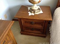 Wood night table with two drawers and metal pulls, placed beside a bed and carpeted floor visible.