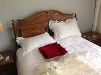 Full bed showing wooden headboard with carved floral details, white eyelet comforter, pillows, and red throw blanket.