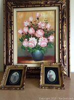 Full view of the framed oil painting of pink roses in a blue vase with two smaller framed prints in front.