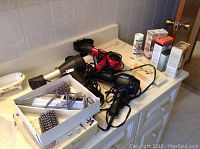 Photo showing multiple hair grooming tools including Braun volume shaper, Conair hair blower, Revlon curling iron, Philips Ultralight 1500 hair dryer, hair brushes, and some boxes on a white surface near a sink.