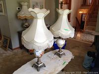 Two vintage table lamps on a table. Each has a glass base with painted figures and metal bases, topped with tall cream shades with gold trim and fringe. Lamps approximately 41 inches tall.