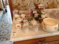 Overview of porcelain and ceramic items on kitchen counter, including JL Menau tea cups, saucers, lidded sugar bowl, creamer, floral mugs, ceramic cups, large bowl, and two decorative battery-operated lights.