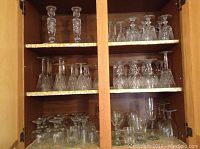 Three shelves in a wooden cabinet filled with assorted cut glass stemware including wine glasses and goblets.