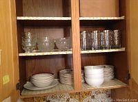 Two shelves with clear Joshua Maxwell bowls, engraved 25th Anniversary glasses, and stacks of white plates in cupboards