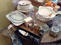 Overview of assortment including white Corning French White casserole dishes, plates, platters, clear casserole dishes, and boxed kitchen items on a table.