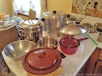 Full overview of cookware including pots, lids, mixing bowls, glass saucepan, and enamel roaster pot on table.