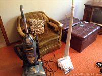 Photo showing Dirt Devil vacuum cleaner upright beside an armchair on orange carpet.