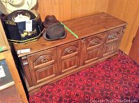 Huppe oak cedar chest shown from a frontal angle with closed panels and metal handles. The chest is set against wood panel wall and red patterned carpet. Several hats and a Dobbs hat box are placed on top.