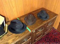 Three men's hats displayed on a wooden surface: a black Dobbs felt hat, a dark brown worn Dobbs felt hat, and a blue felt hat.