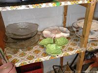 Photo of various glass and china serving ware items on a wooden shelf, including clear glass plates, divided serving trays, and a green leaf-shaped dish.