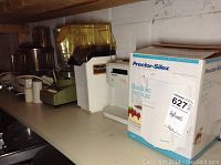 Various kitchen appliances on a shelf including boxed Proctor Silex electric kettle, popcorn maker, hand mixer, and food processor