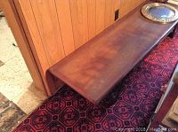 Long low wooden table next to a wood-paneled wall and red patterned carpet, showing surface wear and scratches.