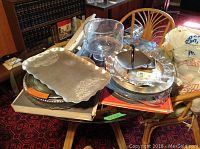 Photo showing an assortment of glass and metal serving trays stacked on a wooden table, including a rectangular aluminum tray with floral embossing and clear glass cake plate with dome cover.