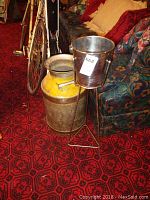 Large metal Teeswater milk can with yellow paint and metal wine cooler bucket on metal stand next to a sofa and bicycle indoors.