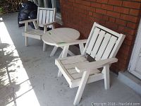 Two white painted wooden patio chairs and a round painted wooden table arranged on a concrete porch with brick wall background. Visible wear includes paint chips and scratches.