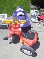 Little Tikes red plastic trike with yellow handlebars and black seat next to a purple Riva Sport plastic sled on pavement with grass background.