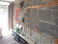 Long view of garden tools hanging on wood rail attached to cement block wall in garage. Tools include shovels, hoes, rake, cultivator, sprinkler, extension cord, watering can, four milk crates, and beige tiles stacked on floor.