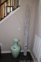 Full view of the mint green vase next to the tall bundle of white decorative reeds in entryway corner.