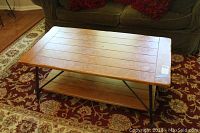Full view of rectangular wooden coffee table with metal legs and bottom shelf placed on a red patterned rug in front of a green sofa.