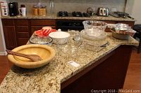 Photo of all serving and bakeware items arranged on a kitchen countertop: wooden bowl, glass bowls, ceramic covered casserole, metal cake pans, aluminum backing pan, and glass platter items.