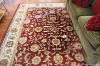 Full view of the area rug showing red floral pattern with ivory border and red trim alongside a green couch with red pillows.