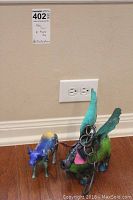 Green and blue metal flying pig next to blue and yellow ceramic cow on hardwood floor near wall outlet